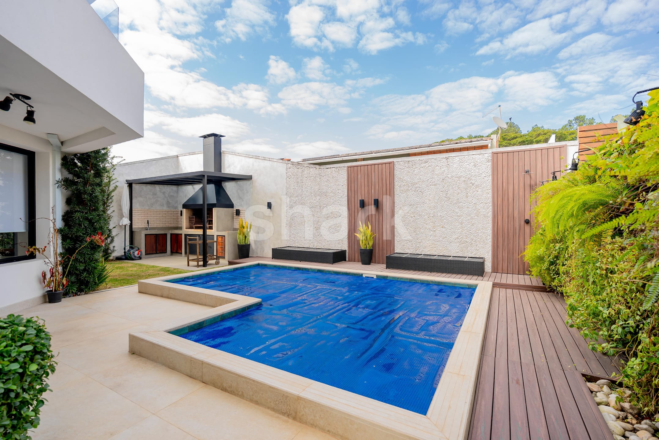 Piscina com deck de madeira e churrasqueira coberta por pergolado ao fundo